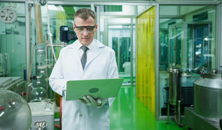 Technician in lab coat and safety glasses recording data on a laptop in a clean, controlled lab environment